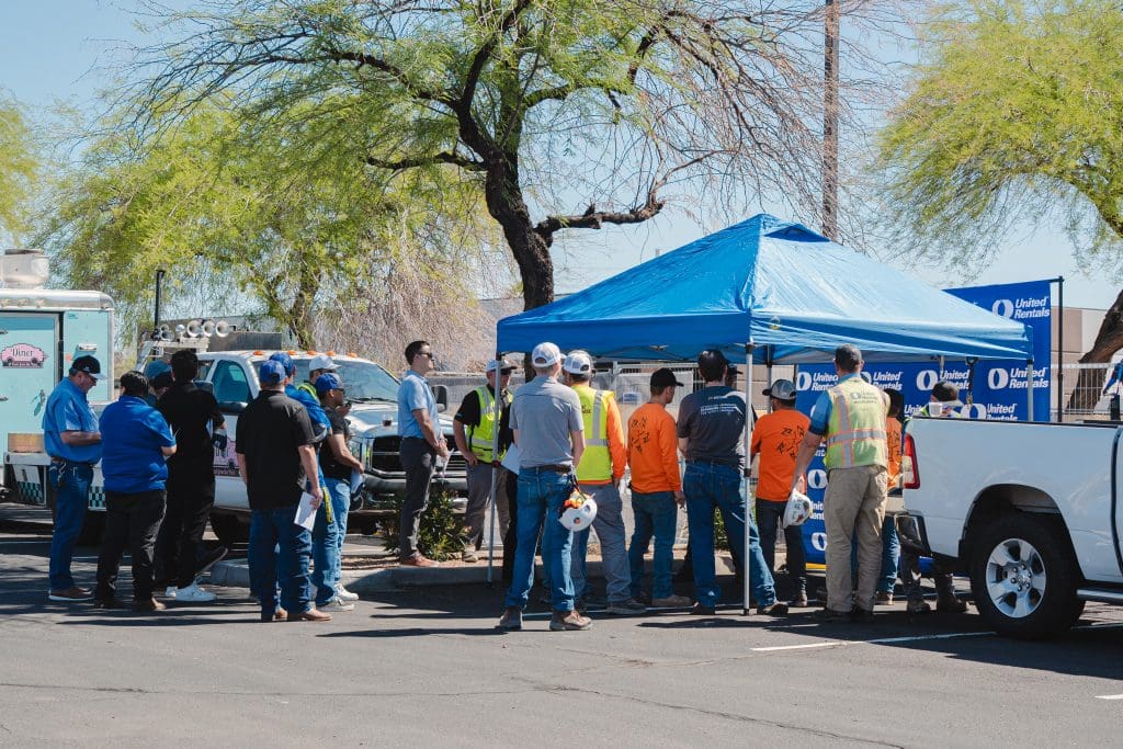 Construction workers and safety personnel gather under a blue tent for a Mobile Elevating Work Platform (MEWP) safety training during Construction Safety Week 2025 in Phoenix, AZ